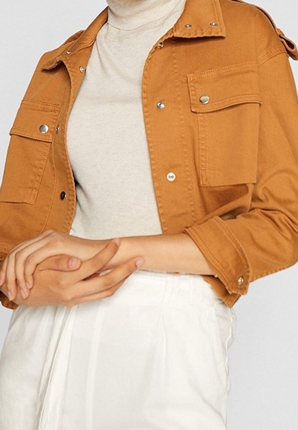 Brun denimjakke med sølvfarvede trykknapper, to brystlommer og en afslappet pasform. Båret over en lys beige turtleneck.