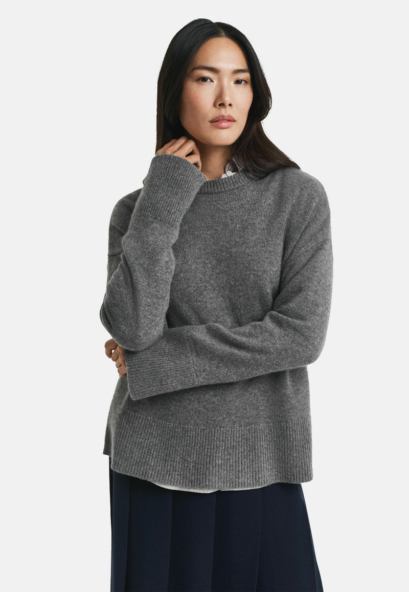 Grauer Strickpullover mit langen Ärmeln, rundem Ausschnitt und geripptem Saum. Das Material wirkt weich und texturiert, ideal für kühlere Wetterbedingungen.