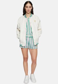 Giubbotto bomber bianco sopra una camicia a righe multicolore e pantaloni corti, con righe verdi, viola e bianche. Sneakers bianche completano l'outfit.