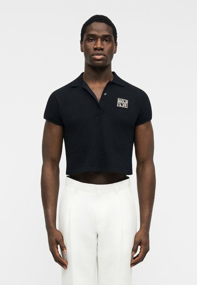 Homme mince portant un polo noir court avec un logo blanc et un pantalon blanc taille haute, debout devant un fond uni.