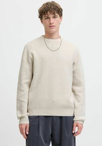 Cremfarbener gestrickter Pullover mit Rundhalsausschnitt, langen Ärmeln und gerippten Bündchen, getragen über dunkelgrauen Hosen. Silberschmuckdetails.