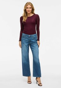 Bordeauxrode lange mouwen top met een textuurpatroon, gecombineerd met blauwe spijkerjeans met wijde pijpen. Model draagt zwarte hoge hakken.