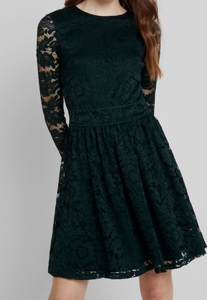 Vrouw draagt een zwarte kanten jurk tot op de knie met lange doorschijnende kanten mouwen en een aansluitende taille tegen een grijze achtergrond.