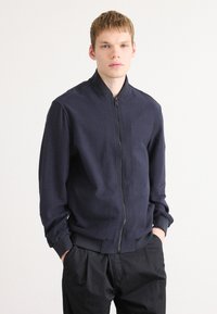 Jonge man met kort blond haar, gekleed in een marineblauwe ritsjack en zwarte broek, met de handen in de zakken, tegen een effen lichte achtergrond.