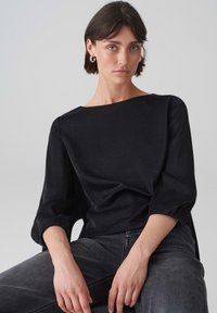 Schwarze, strukturierte Bluse mit weitem Halsausschnitt, 3/4-langen Puffärmeln und lockerer Passform; getragen mit dunkelgrauen Jeans.