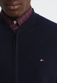 Marinblå zip-up cardigan med strukturerat stickat tyg. Har en hög krage och en liten broderad logotyp i röd, vit och blå. Rutigt skjorte under.