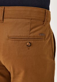 Braune Baumwollhosen mit glatter Textur, ausgestattet mit einer Rückentasche, die mit einem Knopf gesichert ist, und einem kleinen Markenetikett.