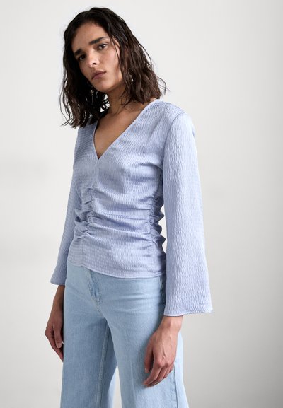 Lyseblå topp med v-hals, rynket front og lange klokkeermer, laget av teksturert stoff. Kombinert med lyse denimbukser.