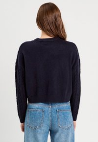 Granatowy dziergany sweter crop z plecionymi detalami wzdłuż rękawów i ściągaczem u dołu. Noszony z jasnoniebieskimi spodniami o wysokim stanie.