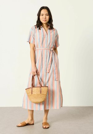 Frau, die ein knielanges, kurzärmeliges, gestreiftes Kleid mit Stoffgürtel trägt, eine geflochtene Handtasche hält und braune Sandalen anhat.