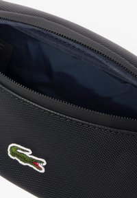 Crna torbica s teksturiranom vanjštinom i glatkim patentnim zatvaračem. Unutrašnjost ima tamnoplavu podstavu. Lacoste krokodil logo ušiven je sprijeda.