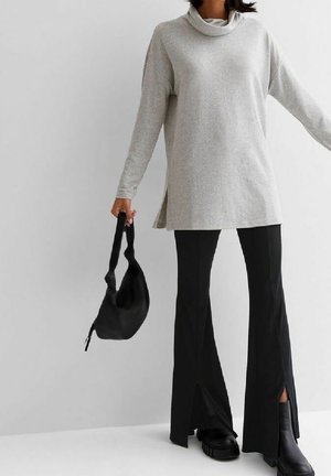 Graue Tunika mit Kapuze und seitlichen Schlitzen, kombiniert mit schwarzen, ausgestellten Hosen mit Schlitzen. Eine schwarze Tasche und klobige schwarze Schuhe runden den Look ab.