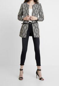 Blazer con motivo bianco e nero, colletto rotondo, vita cinturata e maniche lunghe, abbinato a jeans neri skinny e tacchi a strappi neri.