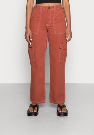 Mujer con pantalones cargo de pierna ancha color óxido y grandes bolsillos delanteros y laterales, combinados con sandalias negras de plataforma.