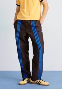 Rjave trenirke z dvema modrima navpičnima črtama, elastičnim pasom in rahlo teksturo. V kombinaciji z rumeno srajco in športnimi čevlji.