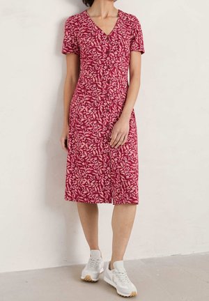 Czerwona sukienka w kwiaty z krótkimi rękawami, zapinana na guziki, z dekoltem w kształcie litery V, sięgająca do kolan, wykonana z lekkiego materiału; zestawiona z białymi butami sportowymi.