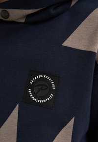 Sudadera de marinero con patrón geométrico beige, con una etiqueta negra que lleva un logo blanco. Fabricada en un suave tejido de punto, de textura lisa.