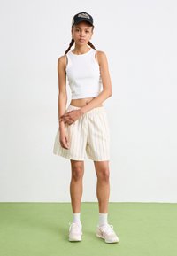 Top cropped branco sem mangas combinado com calções cream com riscas verticais vermelhas. O modelo usa um boné preto e ténis cor-de-rosa com solas brancas.