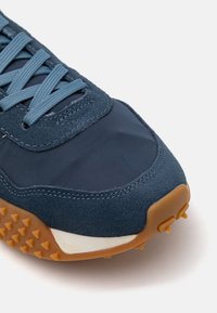 Sötétkék sportcipő, amely velúr és hálós anyagból készült, texturált gumi talppal, apró tüskékkel a tapadásért, valamint kék fűzőkkel.