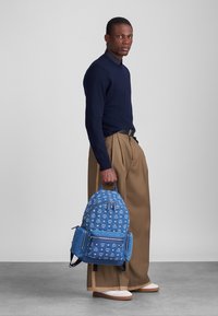 Modrá džínová batoh s bielymi grafickými vzormi, viacerými oddeleniami a nastaviteľnými popruhmi, držený osobou v námorníckom svetri a béžových nohaviciach.