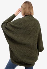 Eine Frau mit blonden Bob-Haarschnitt, die einen übergroßen olivgrünen Strickpullover mit einem strukturierten Rautenmuster trägt und vor einem weißen Hintergrund steht.