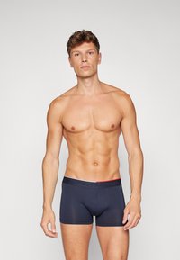 Tumed pokkeri aluspesu, mis sobib keha külge, valmistatud venivast kangast. Omab kontrastset vööd, millel on valge ja punane "TOMMY HILFIGER" bränding.