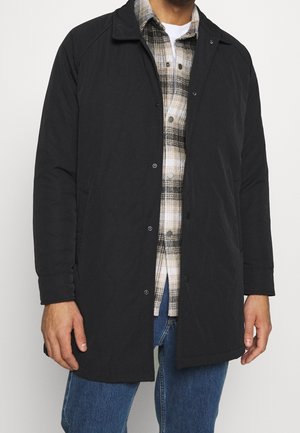 Mężczyzna ubrany w czarny płaszcz na beżową i czarną koszulę w kratę oraz niebieskie dżinsy, stojący na jasnym tle.