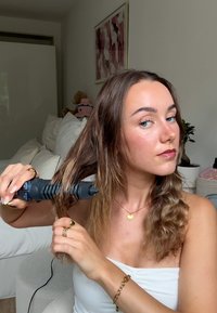 Frau mit hellbraunen Haaren benutzt einen blauen Lockenstab, um ihre Haare in einem Schlafzimmer mit weißen Wänden und Dekor zu stylen.