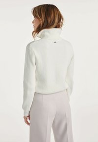 Biały sweter z prążkowanym wzorem i długimi rękawami, z wysokim kołnierzem oraz małą metką marki z tyłu. Połączony z jasnobeżowymi spodniami.