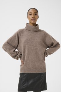 Brown, oversized knitted sweater with a high collar, raglan sleeves, and a ribbed hem. Worn over a black leather skirt.