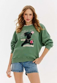Zielona bluza z grafiką psa noszącego perły i beret. Zawiera napisy oraz kwiatowe akcenty. Połączona z dżinsowymi szortami.