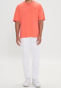 T-shirt a maniche corte di colore corallo, realizzata in tessuto morbido, abbinata a pantaloni bianchi e sneakers bianchi. Branding minimale sulla maglietta.