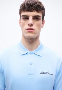 Jeune homme aux cheveux bruns courts portant un polo Lacoste bleu clair, face à l'avant sur un fond blanc uni.