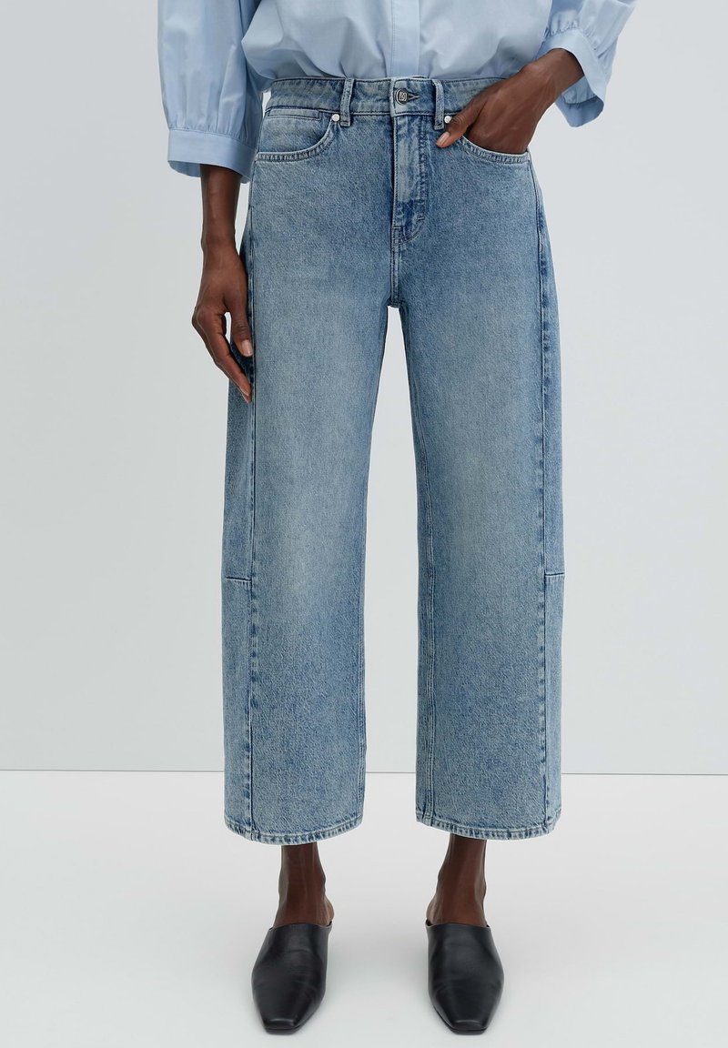 Persona che indossa jeans larghi a gamba larga di colore azzurro chiaro, camicia azzurro chiaro e scarpe nere senza lacci con punta affilata, in piedi su uno sfondo uniforme.