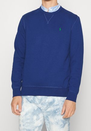 Mand iført blå crewneck-sweatshirt med grønt logo over lyseblå skjorte med knapper og hvide-blå tie-dye bukser, stående mod en hvid baggrund.