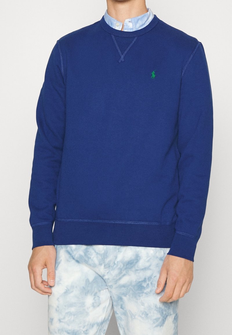 Mann trägt blauen Sweatshirt mit grünem Logo über hellblauem Hemd und weiß-blauen Batik-Hosen, steht vor weißem Hintergrund.