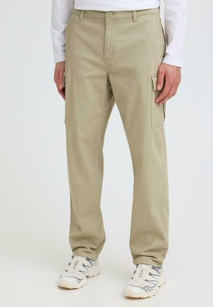 Mann trägt beige Cargo-Hosen mit Seitentaschen und weiße Sportschuhe, steht vor einem schlichten hellen Hintergrund.