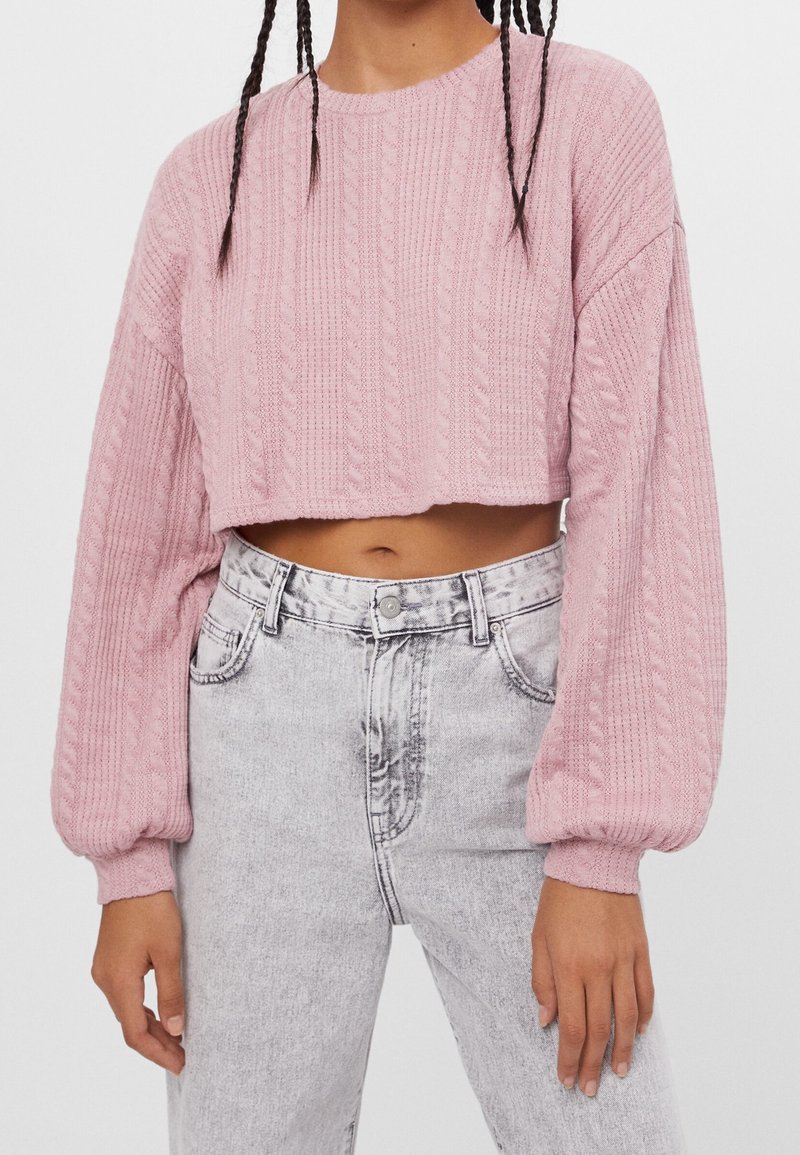 Maglione cropped rosa con motivo a treccia testurizzata, maniche ampie e scollatura rotonda, abbinato a jeans grigi chiari a vita alta.