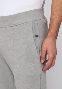 Šedé tepláky z texturované látky, elastický pás, kontrastní šedá boční kapsa s černým knoflíkem, vyšívané logo na stehně.