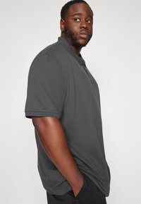 Uomo che indossa una polo grigio scuro e pantaloni neri, con le mani in tasca, mentre guarda oltre la spalla su uno sfondo neutro.