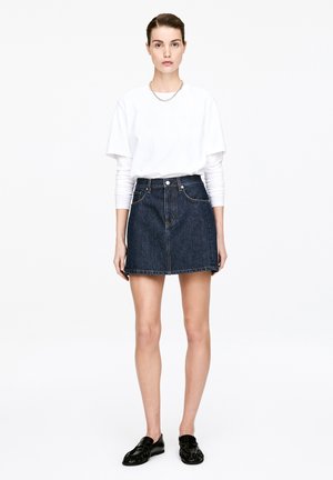 Kobieta stojąca, ubrana w białe koszule z długim i krótkim rękawem, ciemną spódnicę dżinsową, czarne mokasyny oraz srebrny łańcuszek, na tle jednolitego białego tła.