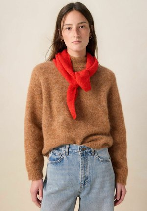 Jeune femme portant un pull marron duveteux, un jean bleu clair et une écharpe rouge vif nouée autour du cou, sur un fond uni.