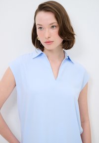 Blusa azzurro chiaro con scollatura a V e maniche a cappuccio corte, realizzata in un materiale morbido e liscio. Presenta un colletto e un dettaglio con un singolo bottone.
