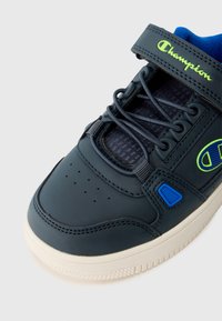 Deportiva azul oscura con una correa de velcro, puntera perforada y lengüeta de tela texturizada. Presenta un logotipo verde y una suela de goma blanca.