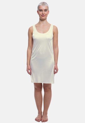 Woman with short blonde hair standing barefoot, wearing a sleeveless pale yellow knee-length dress on a plain white background.