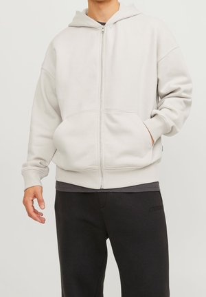 Homme portant un sweat à capuche gris clair avec fermeture éclair, main dans la poche, et pantalon noir, sur un fond clair uni.