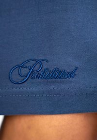 Mørkeblå stof med broderet "Forbudt" tekst i en lysere blå tråd. Glat tekstur med et enkelt design og pæn syning.