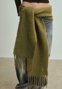 Écharpe en laine vert olive avec une texture douce, aux bords frangés et nouée autour de la taille ; associée à un jean bleu taille haute.