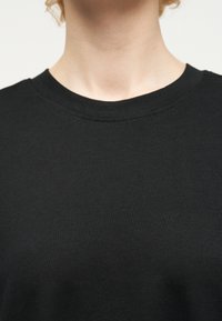 Κοντινή λήψη του λαιμού και του πάνω μέρους του στήθους ενός ατόμου που φοράει απλό μαύρο μπλουζάκι με στρογγυλή λαιμόκοψη, με ουδέτερο φόντο.