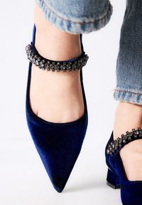 Zapatos de terciopelo azul con punta afilada, tacón grueso y una correa de tobillo adornada con acentos de piedra oscura. Puño de mezclilla visible por encima del zapato.